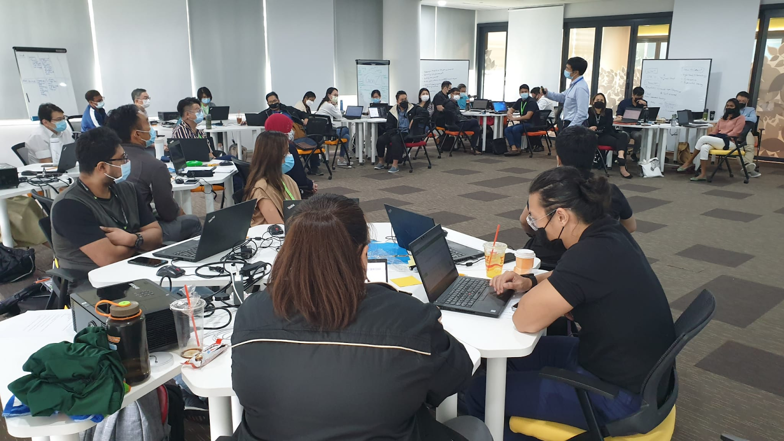 People are sitting in a room with laptops and drinks, listening to a presenter near a whiteboard.